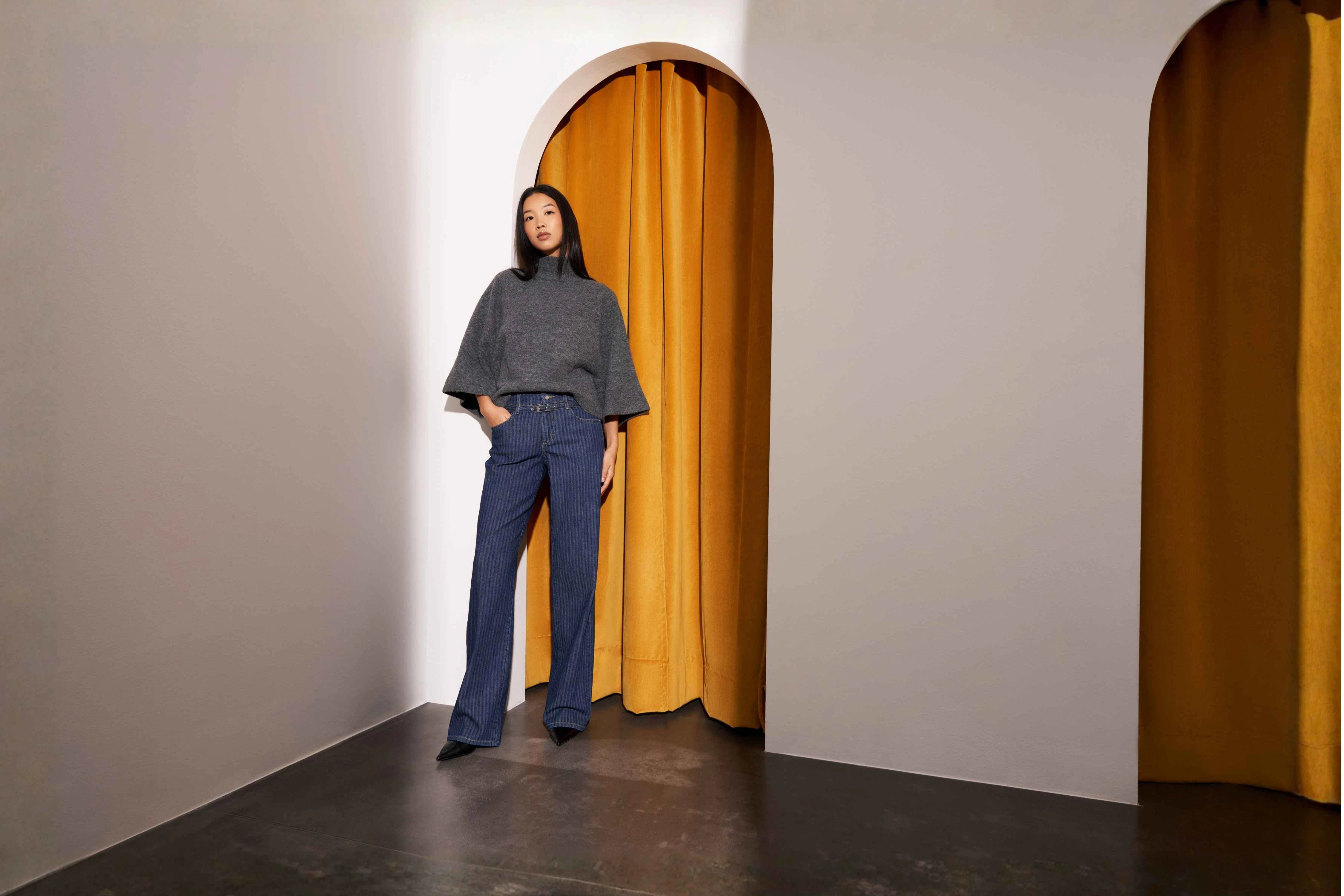 Dunkelhaariges Model trägt eine dunkelblaue Jeans mit hellen Streifen und Gürtel sowie ein graues Oberteil