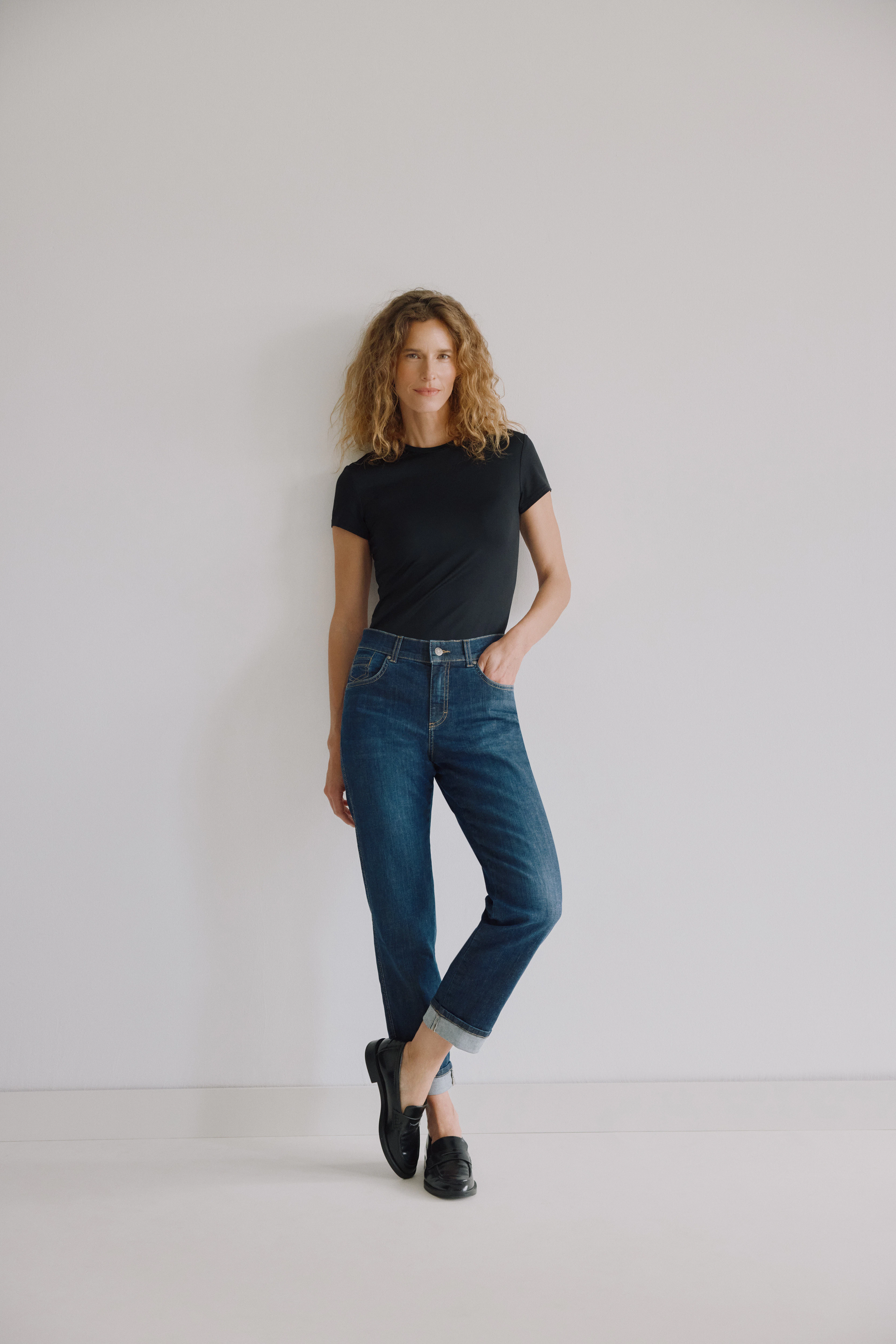 Frau trägt dunkelblaue Cropped Straight Fit Jeans von Angels mit Umschlag, schwarzes T-Shirt und schwarze Loafer