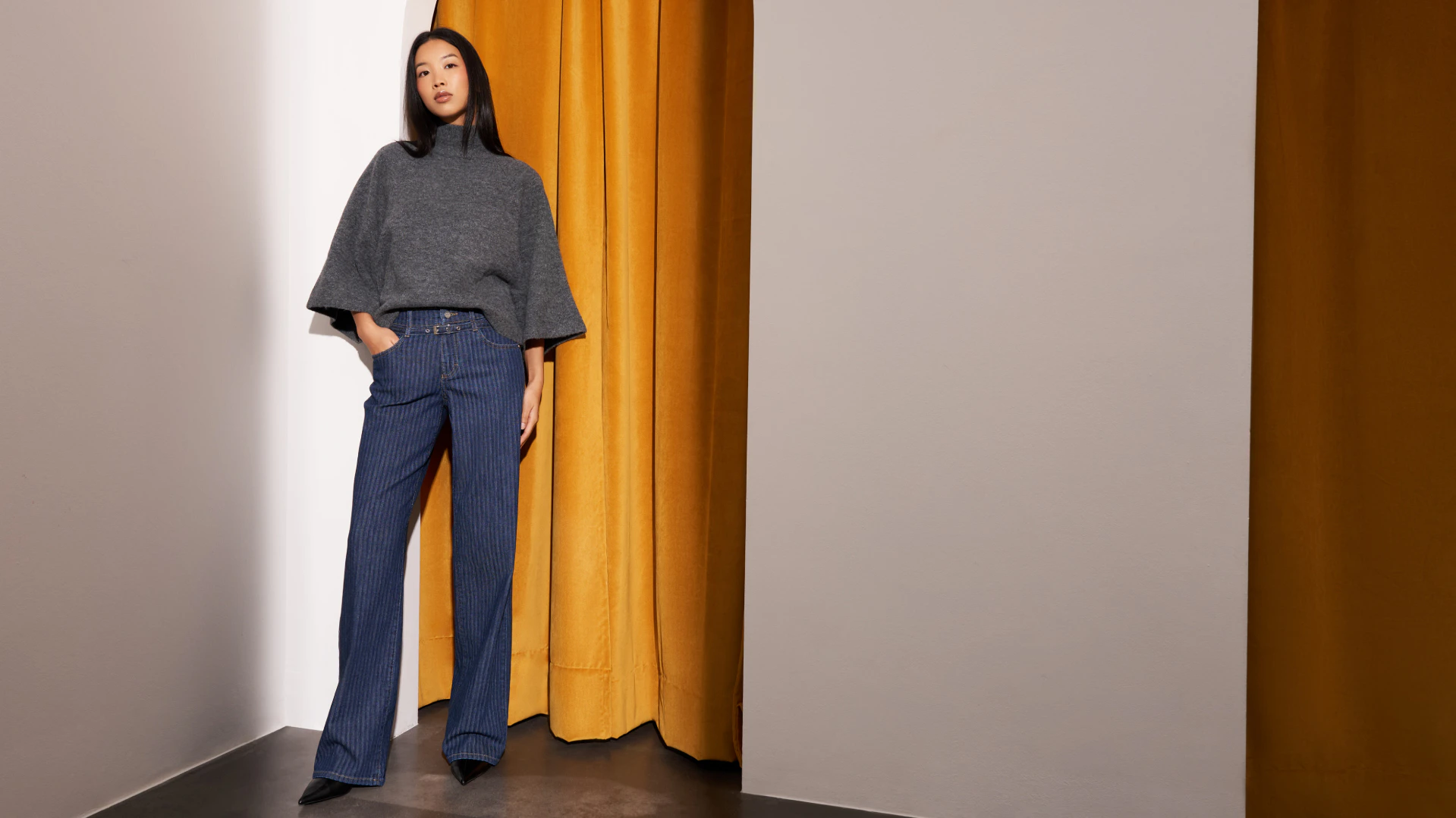 Dunkelhaariges Model trägt eine blaue Jeans mit Streifen und Gürtel sowie ein graues Oberteil.