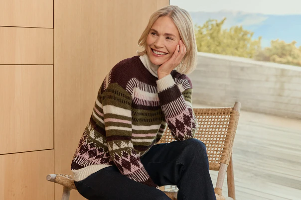 Eine Frau sitzend auf einem Stuhl in blauer Jeans und gemustertem Strickpullover.