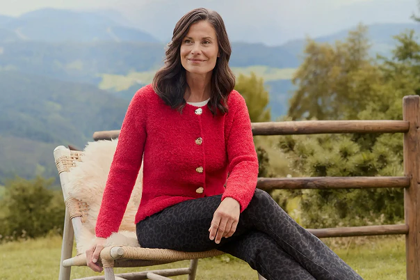 Eine Frau sitzend mit brauner Hose und einer roten Strickjacke.