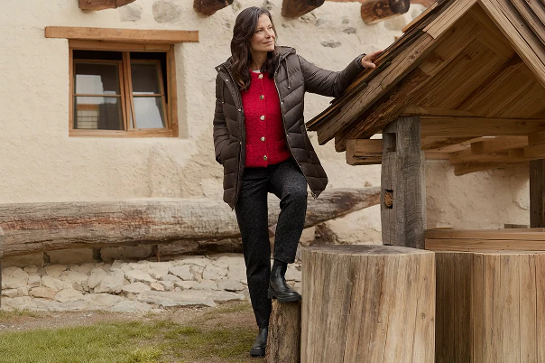 Eine Frau stehend in brauner Hose, roter Strickjacke und brauner Steppjacke. 