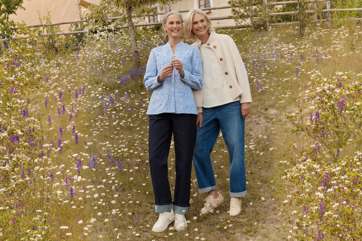 Zwei Frauen stehend in blauer Jeans, blauer Bluse und beiger Jacke. 