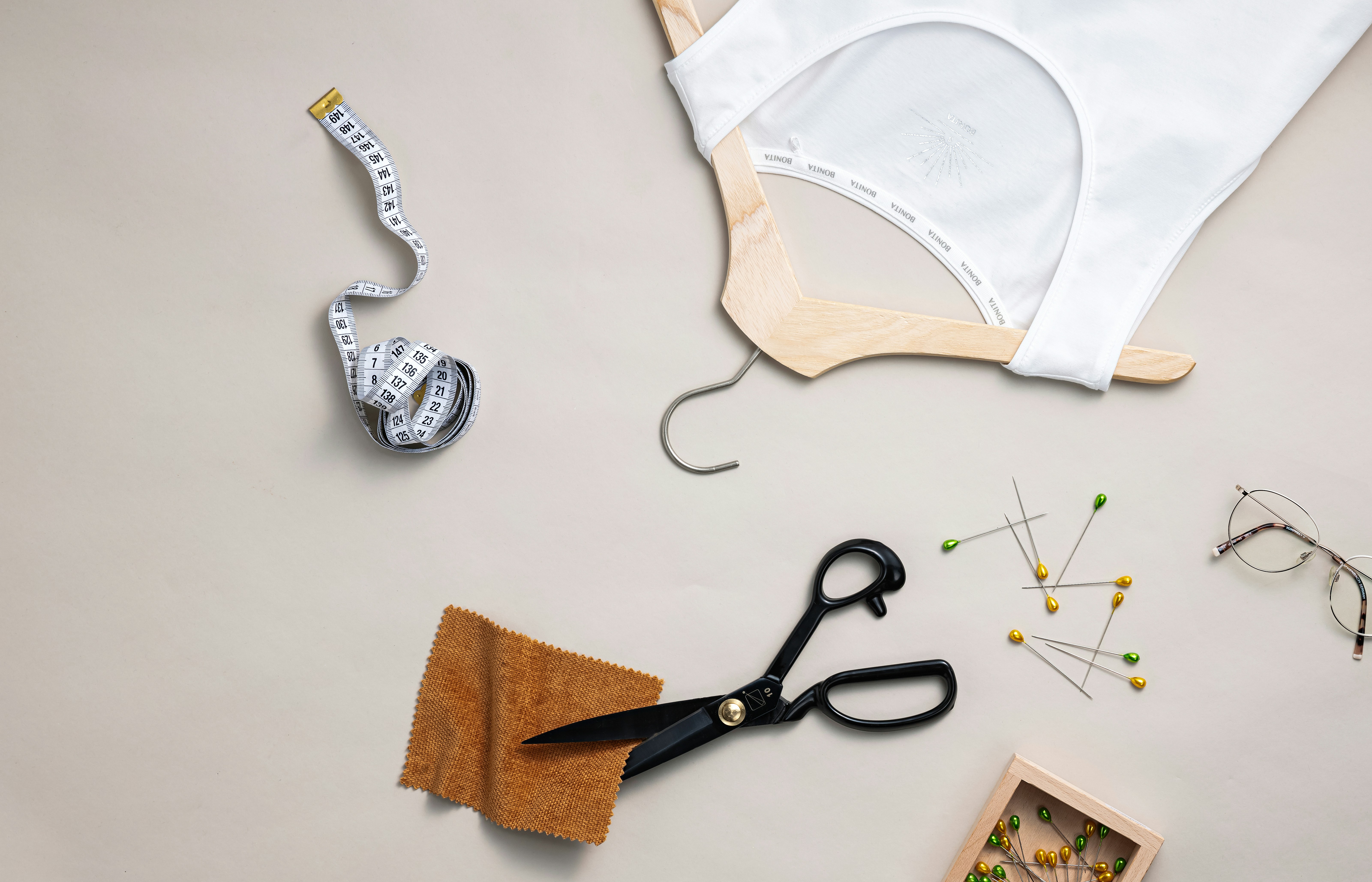 Auf einer hellen Fläche liegen ein weißes Top auf einem Holzkleiderbügel, ein Maßband, eine Schere, ein Stoffmuster, Stecknadeln und eine Brille – Symbole für Modehandwerk und Design.