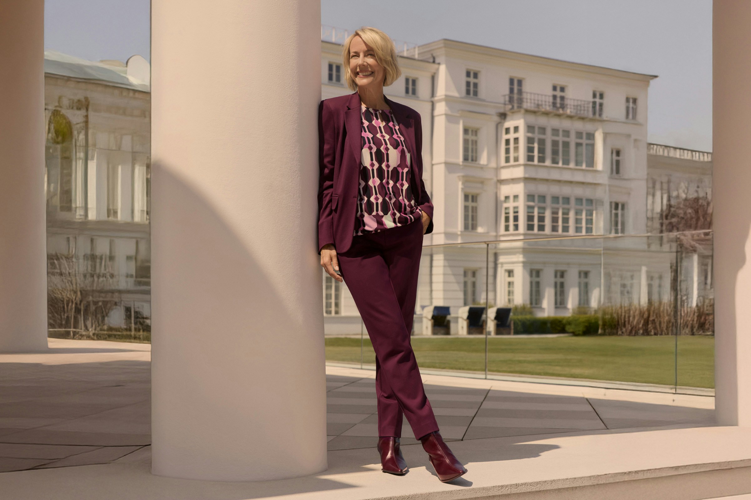 Vrouw in een wijnrood pak leunt glimlachend tegen een zuil voor een groot gebouw.