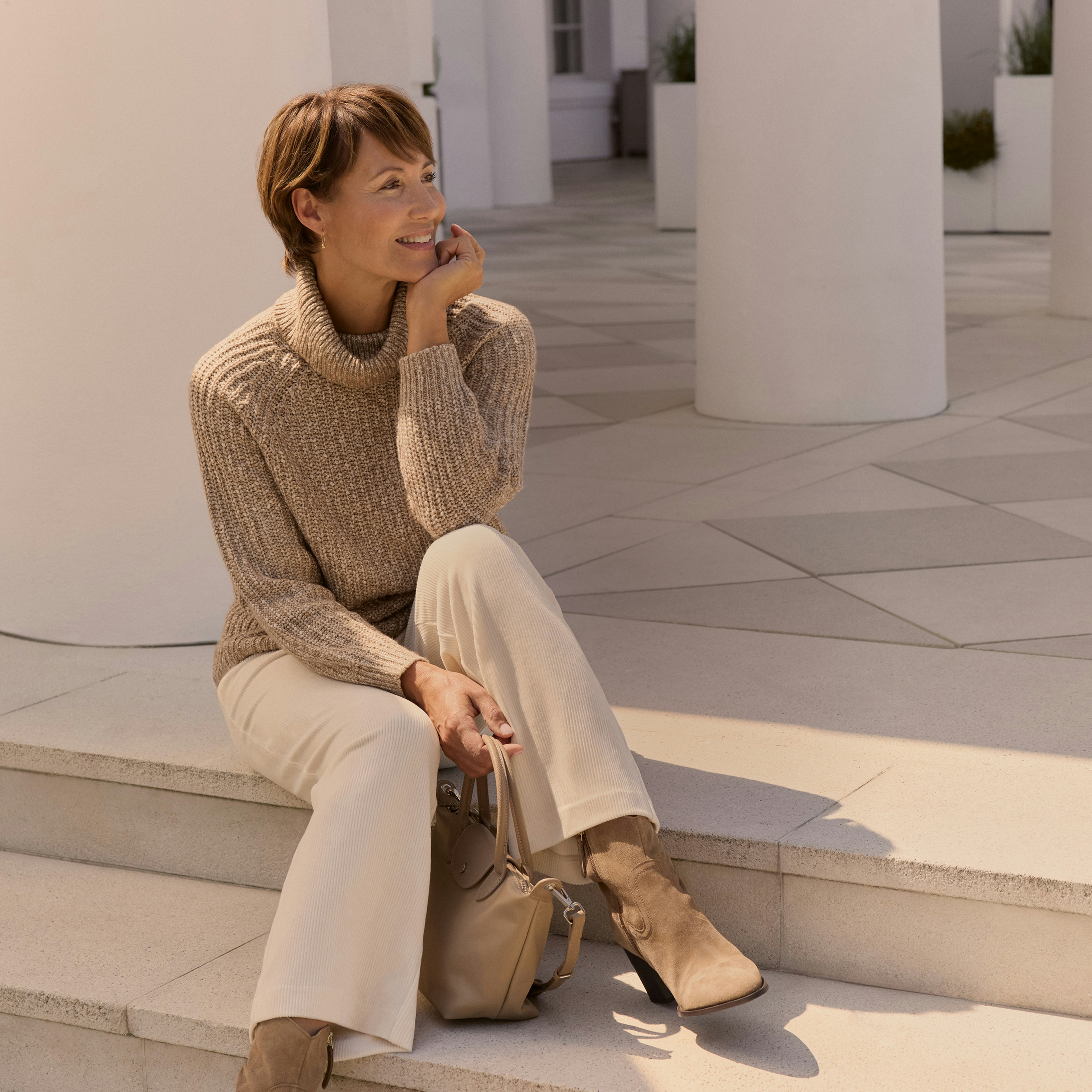 Eine Frau im Bonita Look, sitzend auf einer Treppe in einem kuscheligen Pullover und beiger Hose. 
