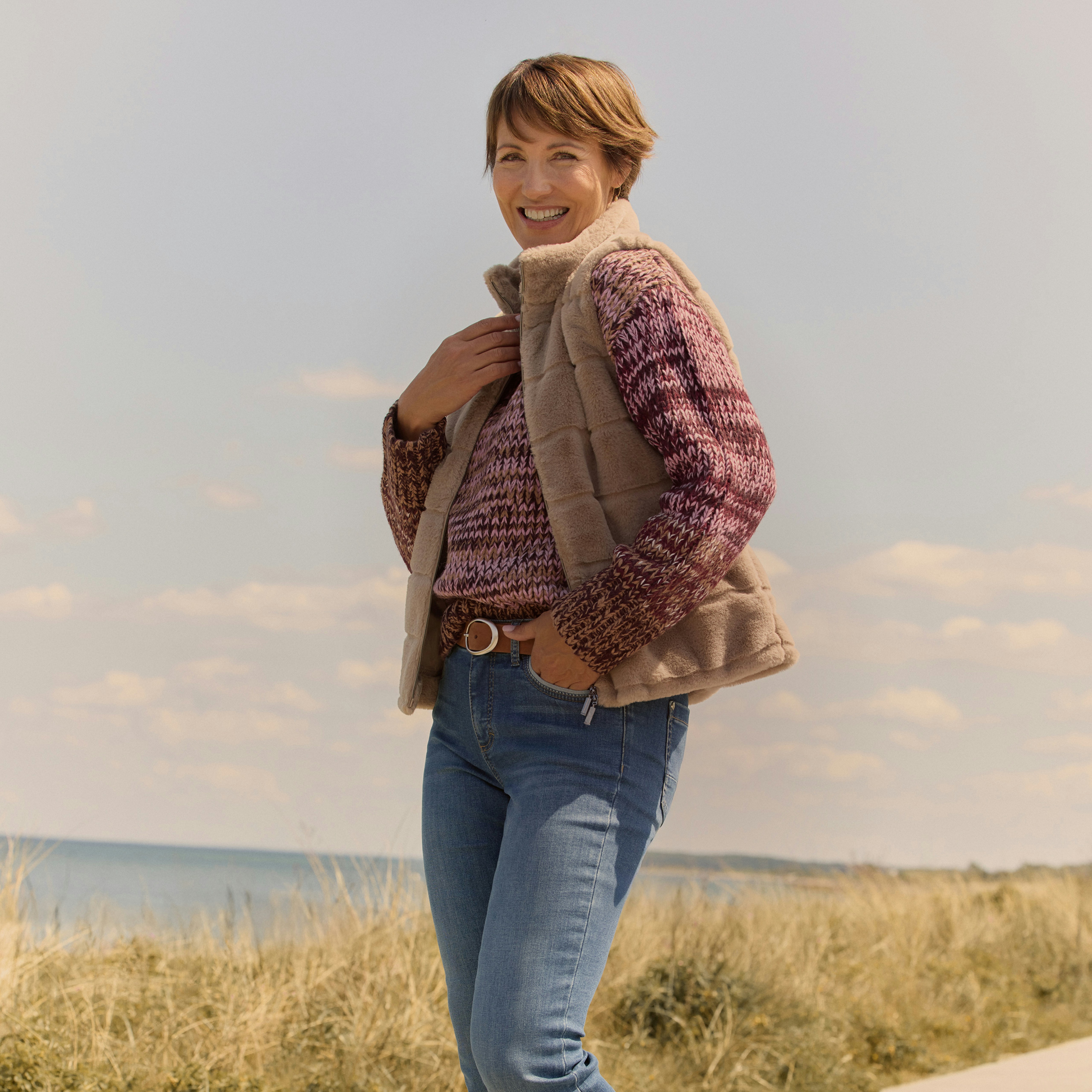 Eine Frau in Bonita Neuheiten, stehend am Strand in blauer Jeans, mit einem kuscheligen Pullover und einer Fellweste. 