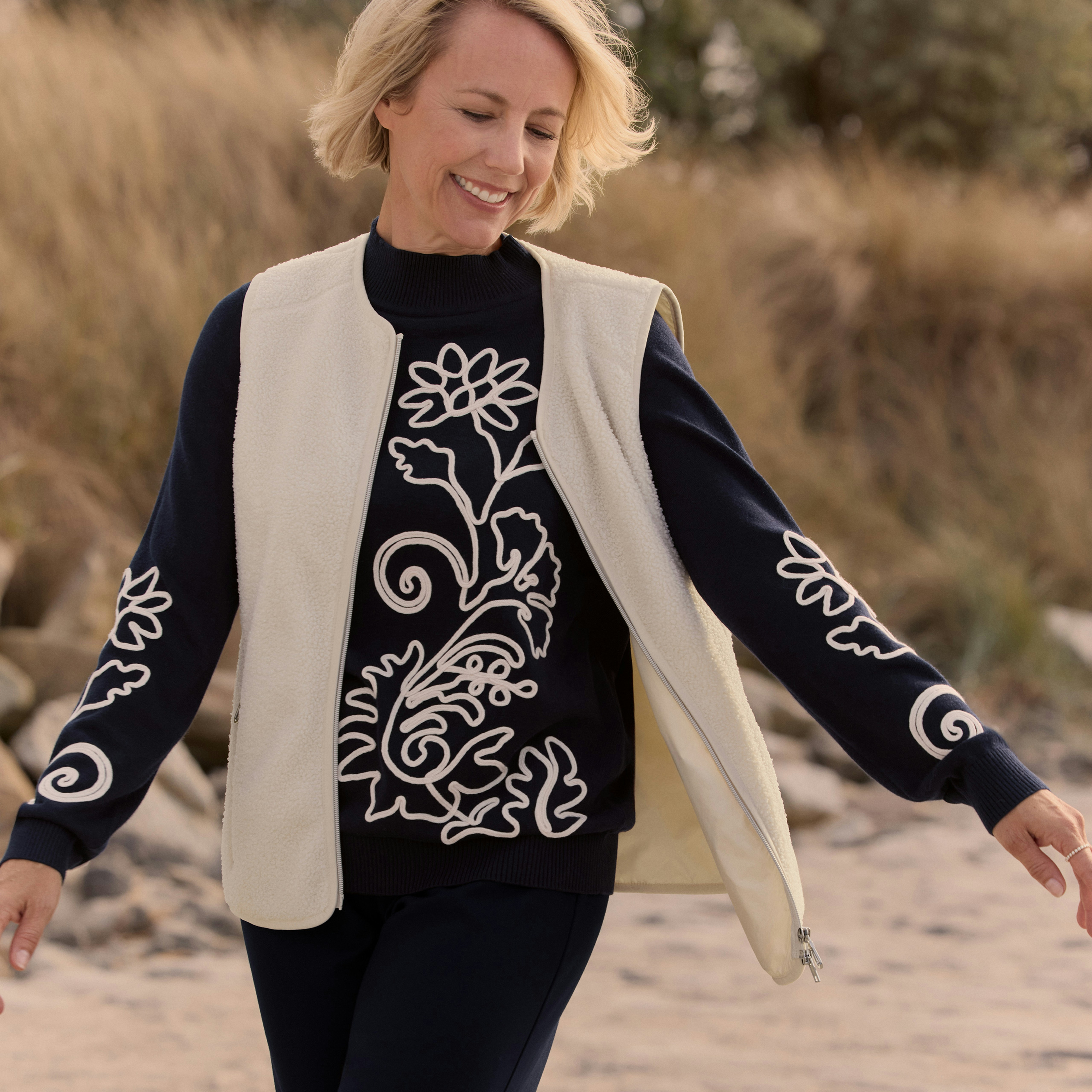 Vrouw draagt de donatie-trui in combinatie met een gebreid vest op het strand. 