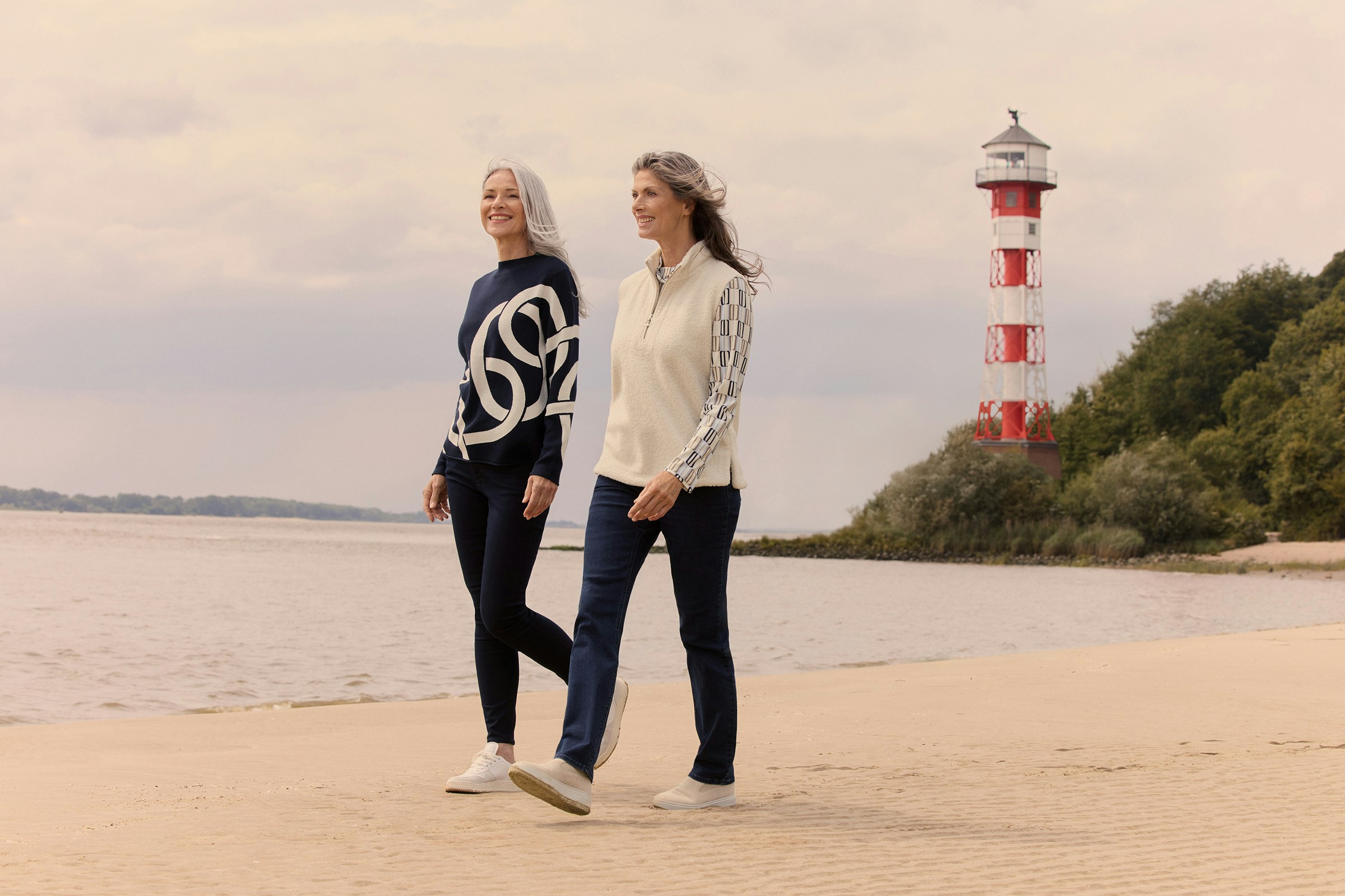 Zwei Frauen mit grauen Haaren spazieren lächelnd am Strand entlang, im Hintergrund steht ein rot-weiß gestreifter Leuchtturm.