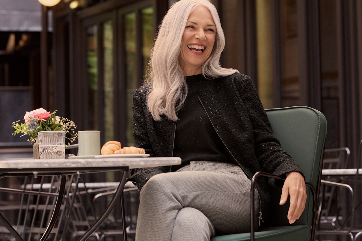 Een vrouw zittend in een grijze broek en een zwart shirt met bijpassend jasje.   