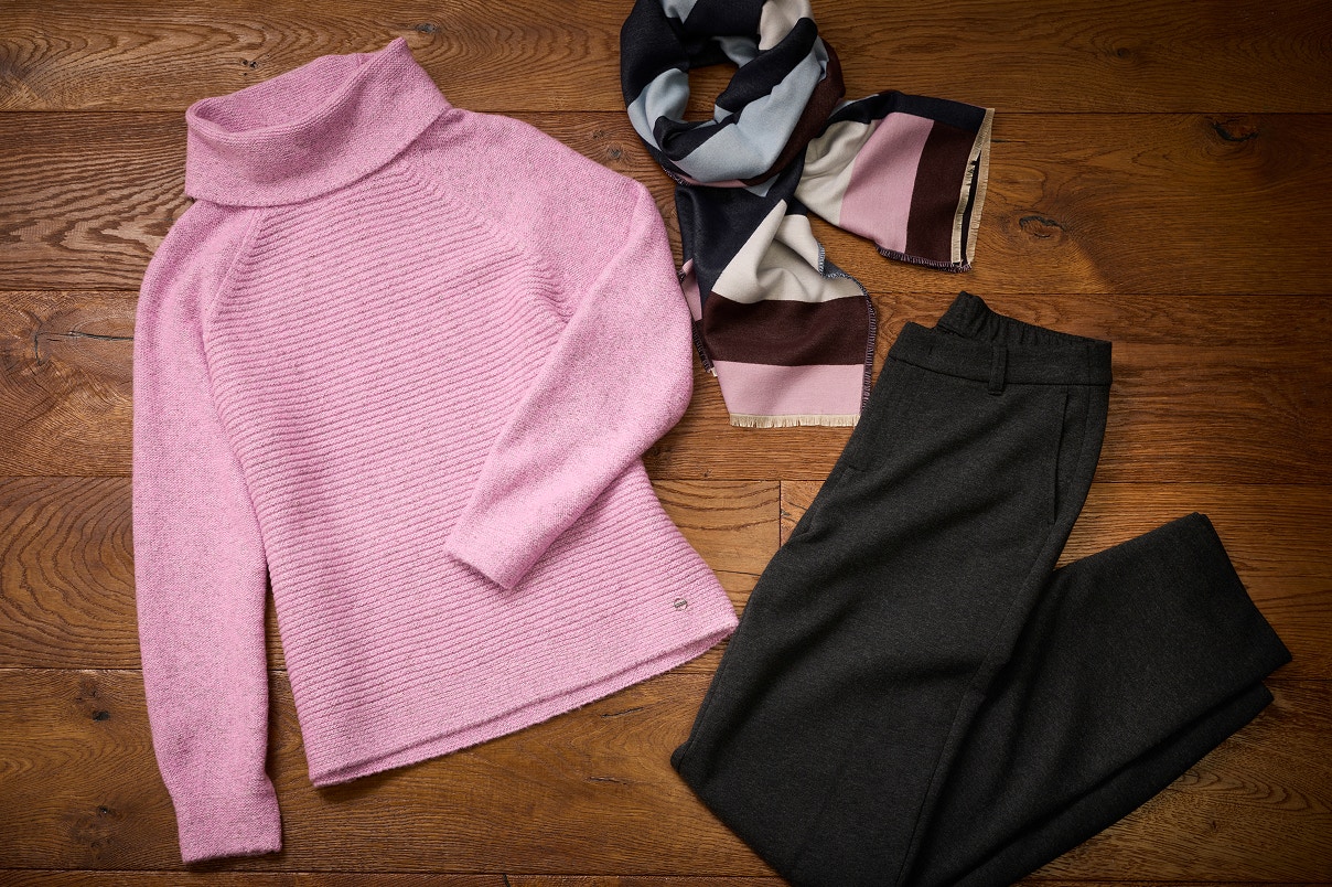 Een outfit liggend op een houten vloer met een roze gebreide trui, een sjaal met patroon en een grijze stoffen broek.