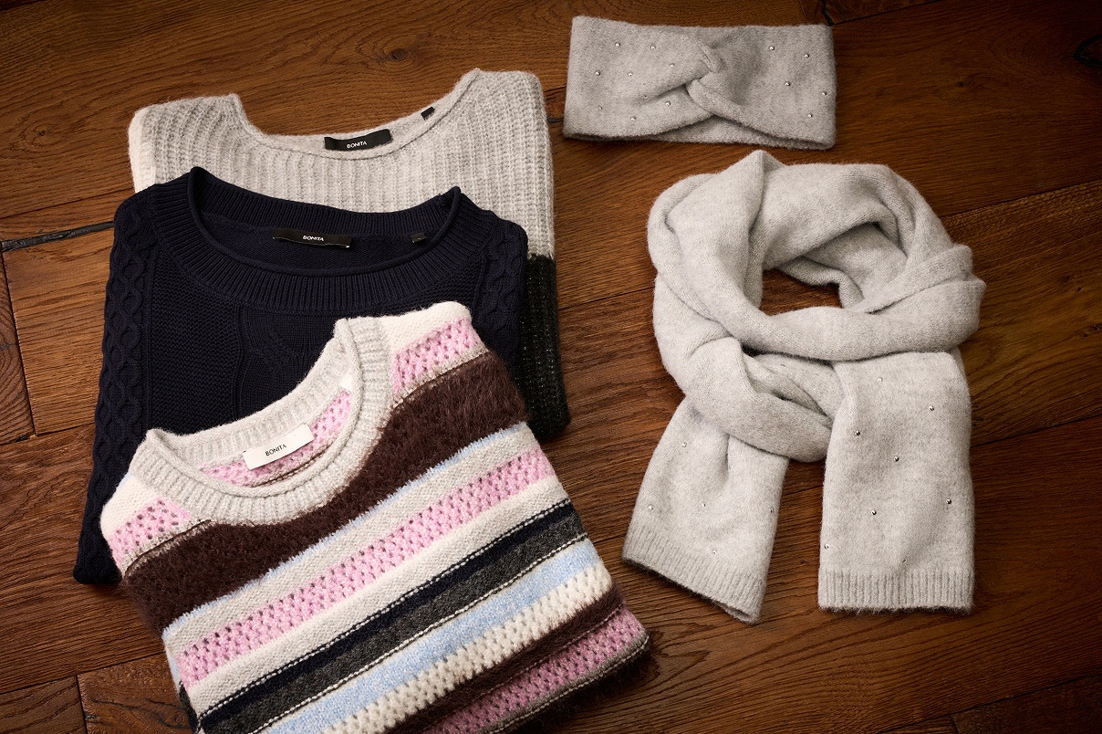 Drie gebreide truien liggen op een houten vloer en een grijze sjaal met bijpassende hoofdband. 