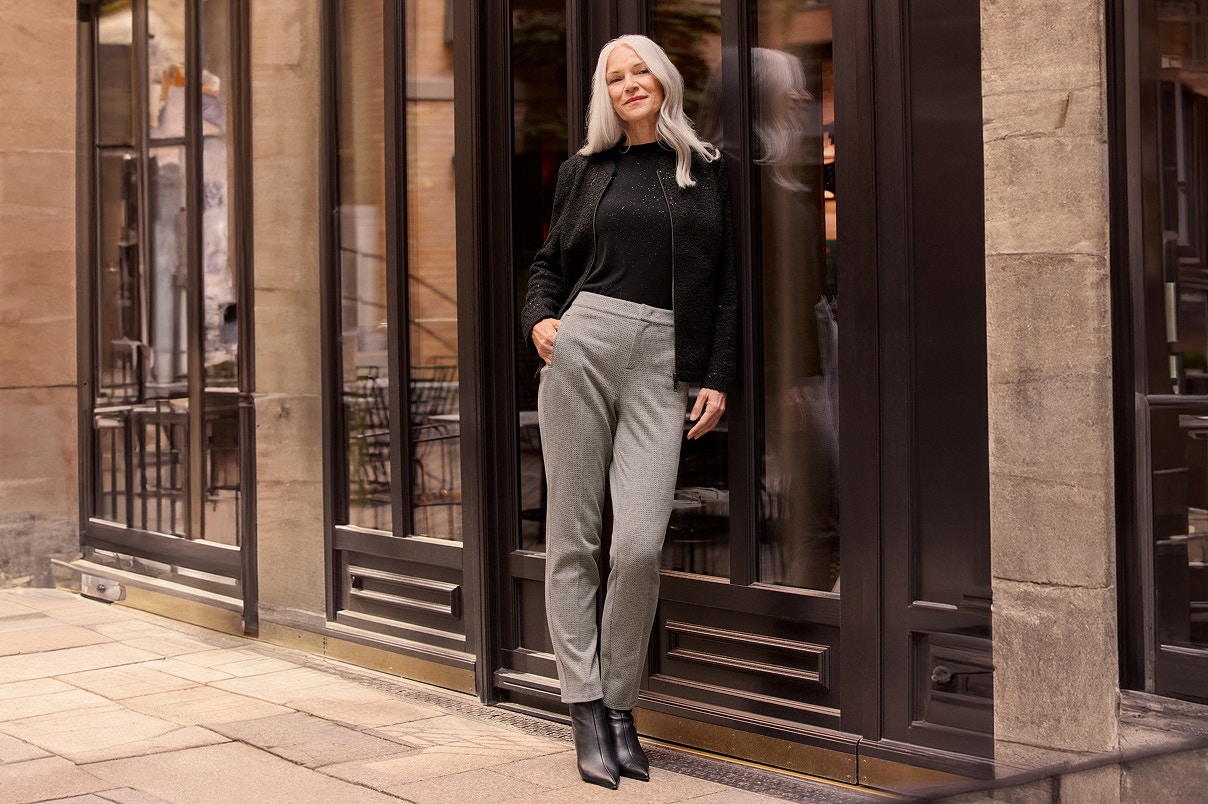 Een vrouw in een grijze broek, met een feestelijk shirt en blazer. 