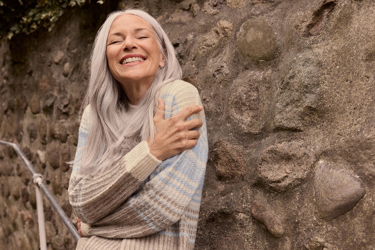 Een vrouw in een Bonita-trui, leunend tegen een stenen muur. 