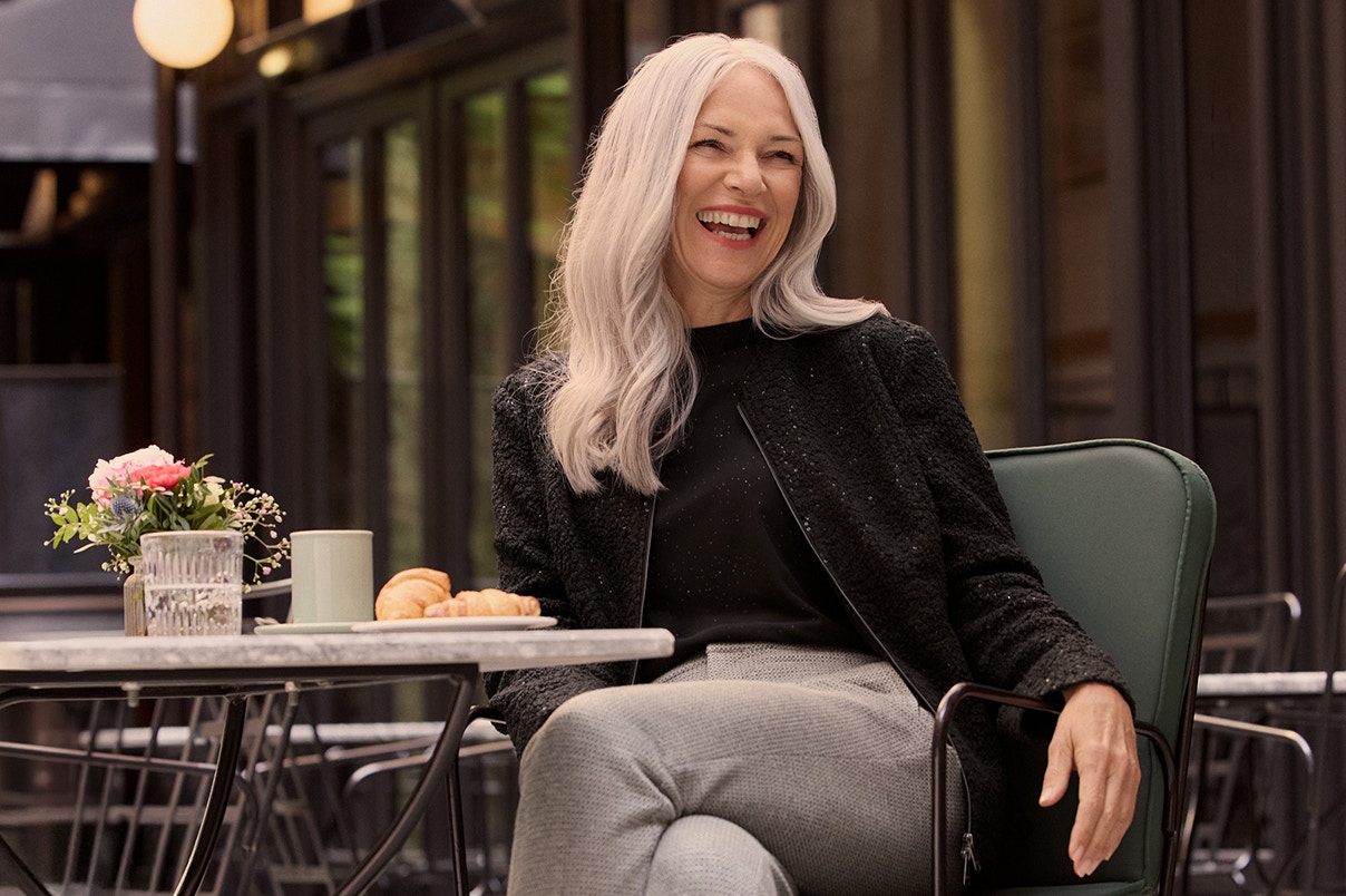 Eine Frau sitzend auf einem Stuhl in einer grauen Hose und schwarzem Oberteil.