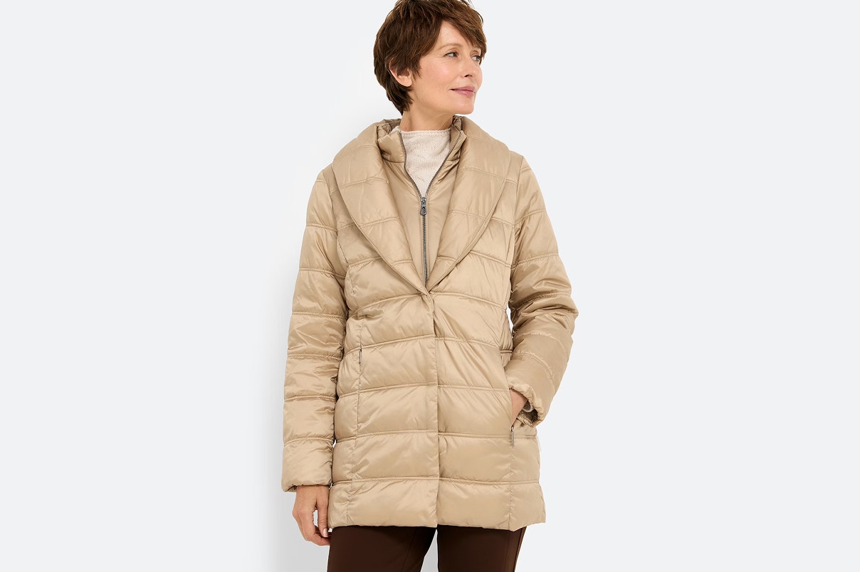 Eine Frau im Bonita Steppmantel, mit beigem Strickpullover und brauner Hose. 