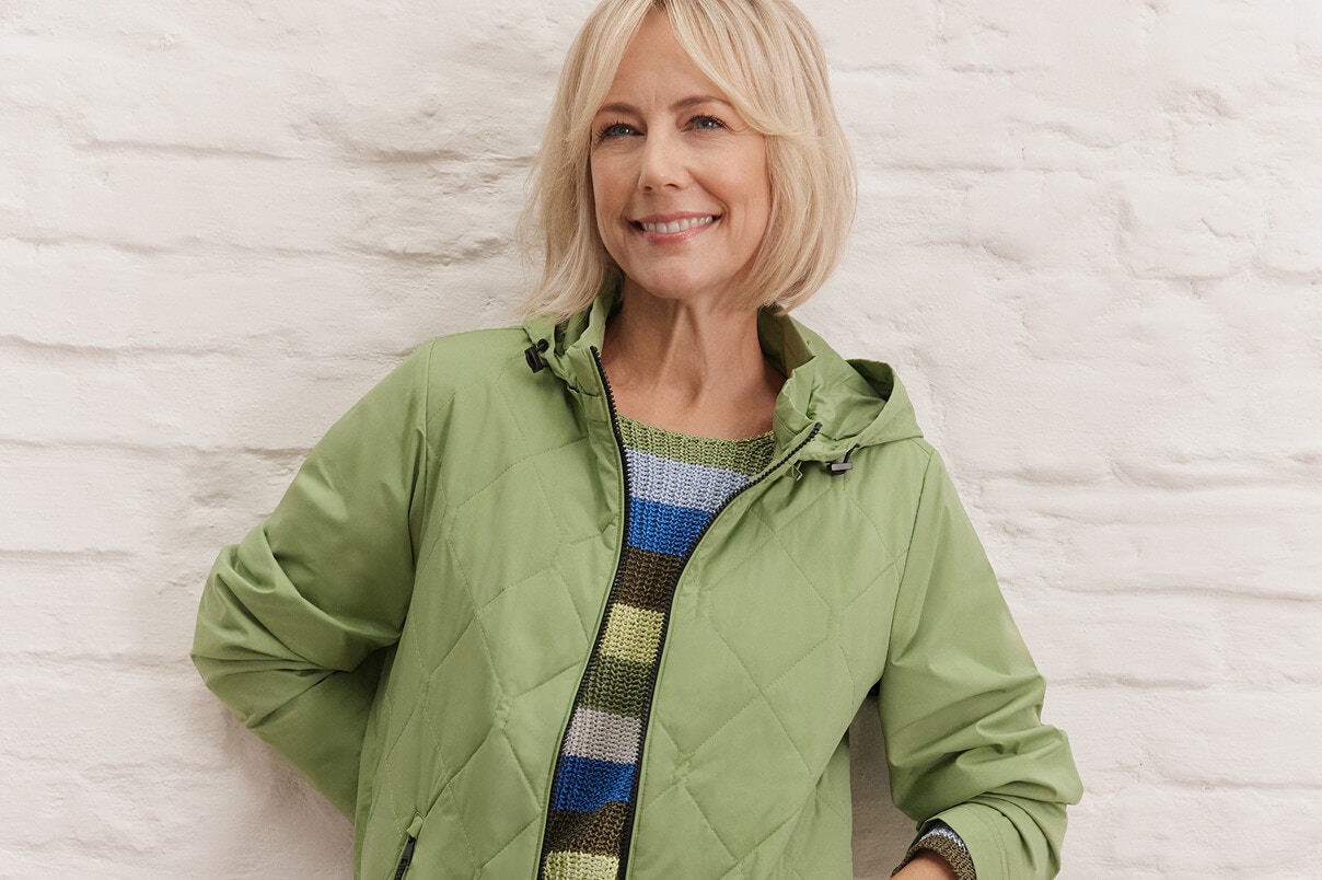 Een vrouw staat in een gestreepte trui, blauwe spijkerbroek en groene jas. 