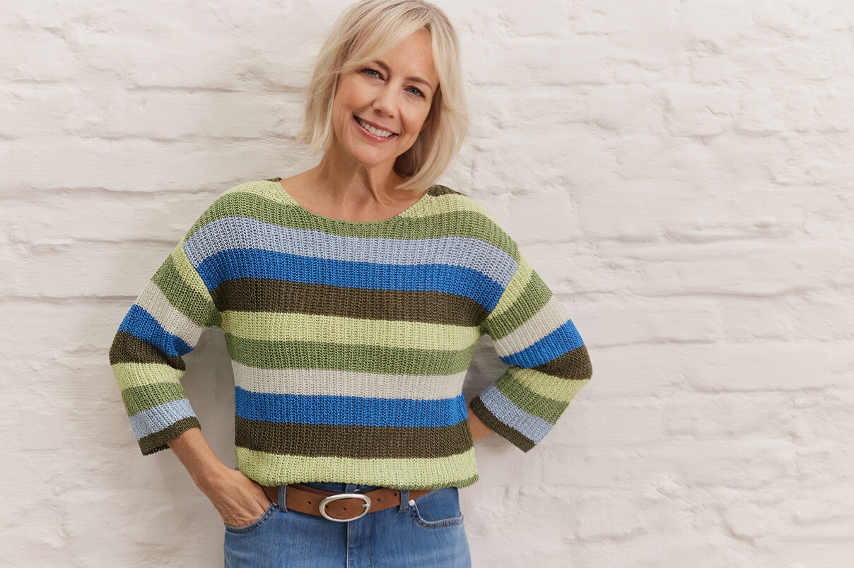 Een vrouw staand in een gestreepte gebreide trui en blauwe spijkerbroek. 