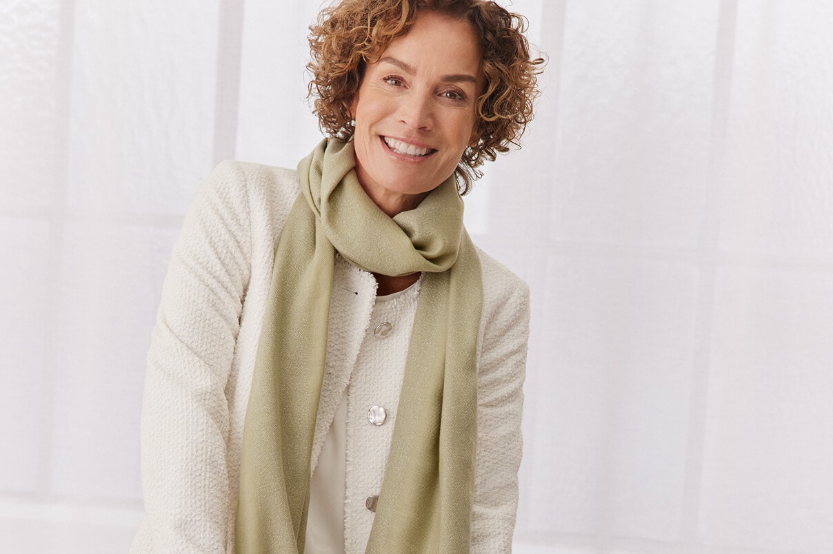 Een vrouw zit op de leuning van een bank in een witte broek, een wit jasje en een groene sjaal.