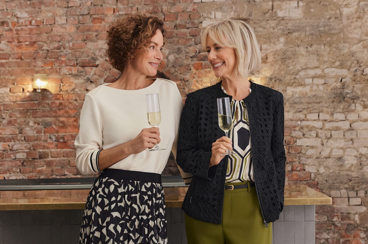 Twee vrouwen staan in Bonita Nieuwigheden, een vrouw in een rok met patroon en een beige top en de tweede vrouw in een groene broek, een top met patroon en een zwart jasje. 