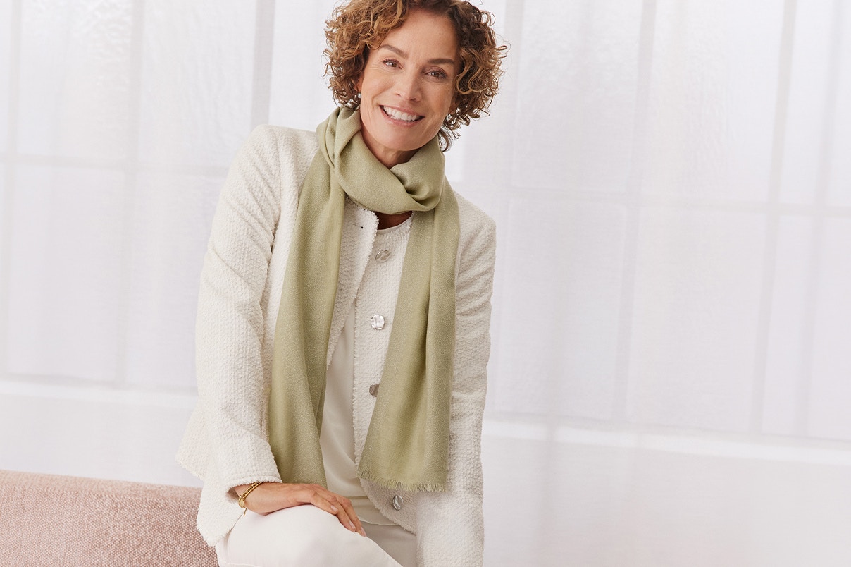 Eine Frau sitzend auf einer Sofalehne in weißer Hose und weißem Blazer und einem grünen Schal. 