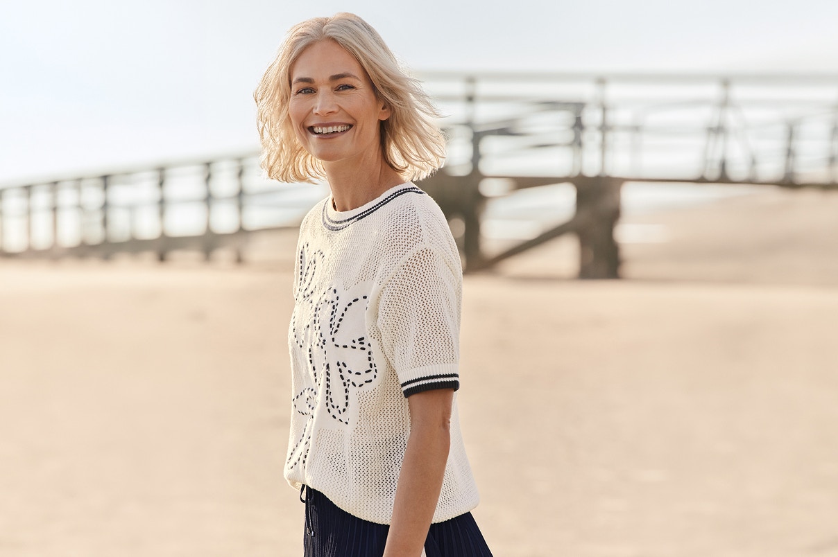 Eine Frau stehend am Strand in einem weißen Shirt und blauem Vorderprint. 