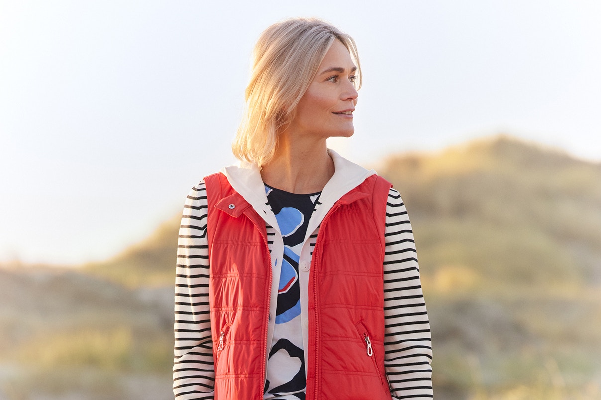 Eine Frau stehend in einem Streifen-Sweatshirt und roter Weste und blauer Hose. 