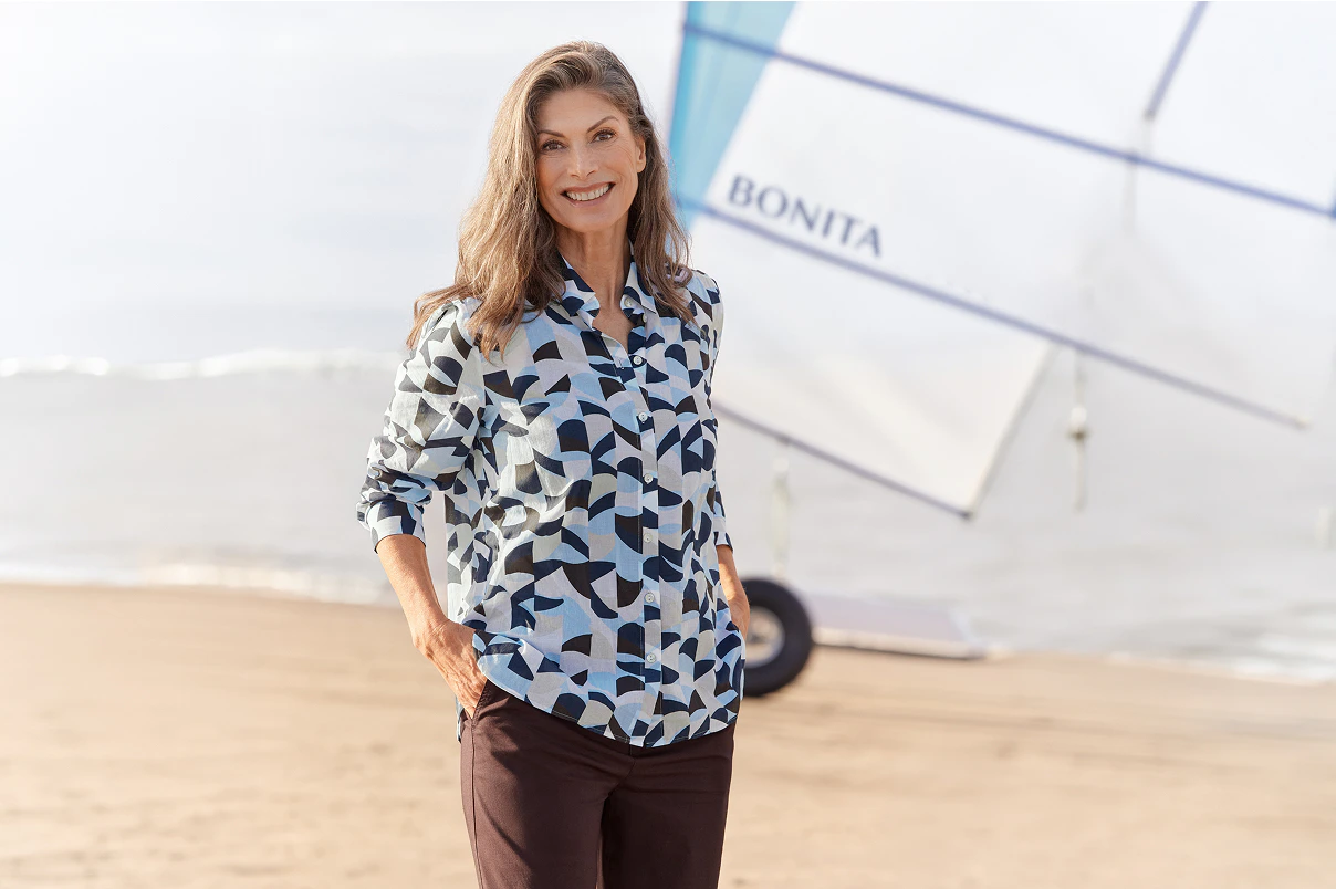 Eine Frau stehend in einer gemusterten Bluse mit brauner Hose.   