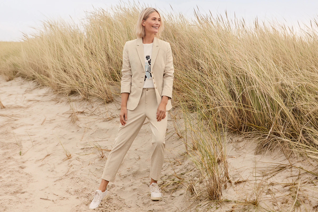 Eine Frau stehend am Strand in einem beigen Hosenanzug und weißem Shirt mit Print. 