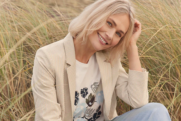 Eine Frau sitzend am Strand in einem weißen Shirt und blauem Vorderprint, Jeansjacke und beiger Hose. 