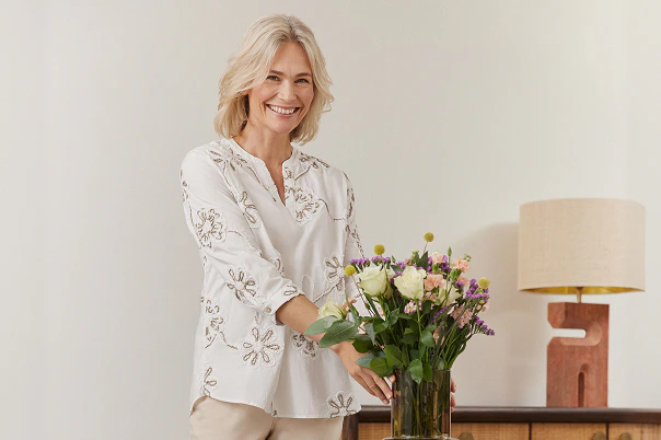 Eine Frau stehend in beiger Hose und weißer Bluse mit Stickereien.  