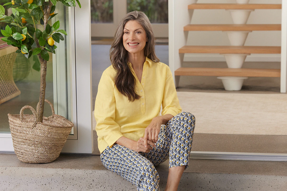 Een vrouw zit op een trede, gekleed in een broek met een patroon en een gele blouse. 