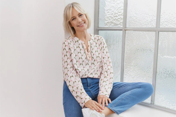 Eine Frau sitzend in blauer Jeans und gemusterter Bluse. 