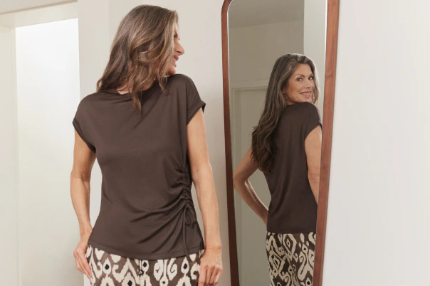 Een vrouw die staat, gekleed in een broek met patroon en een bruin T-shirt.  