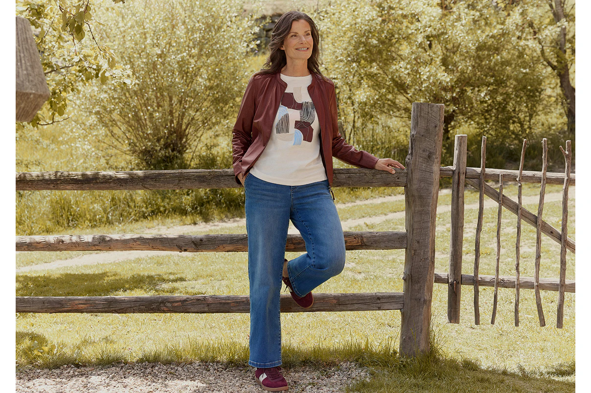 Een vrouw die staat, gekleed in een blauwe spijkerbroek, een T-shirt met print en een imitatieleren jasje.