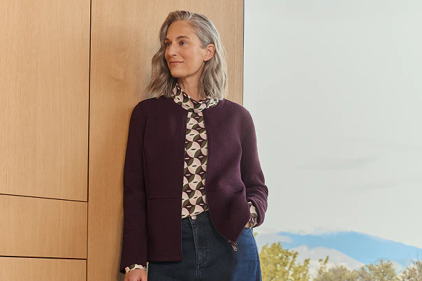 Eine Frau anlehnend an einer Wand in blauer Jeans und gemustertem Shirt und bordeaux Jacke. 