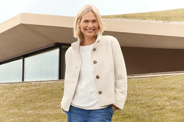 Eine Frau stehend in blauer Jeans und hellem Shirt und kuscheliger Jacke.  
