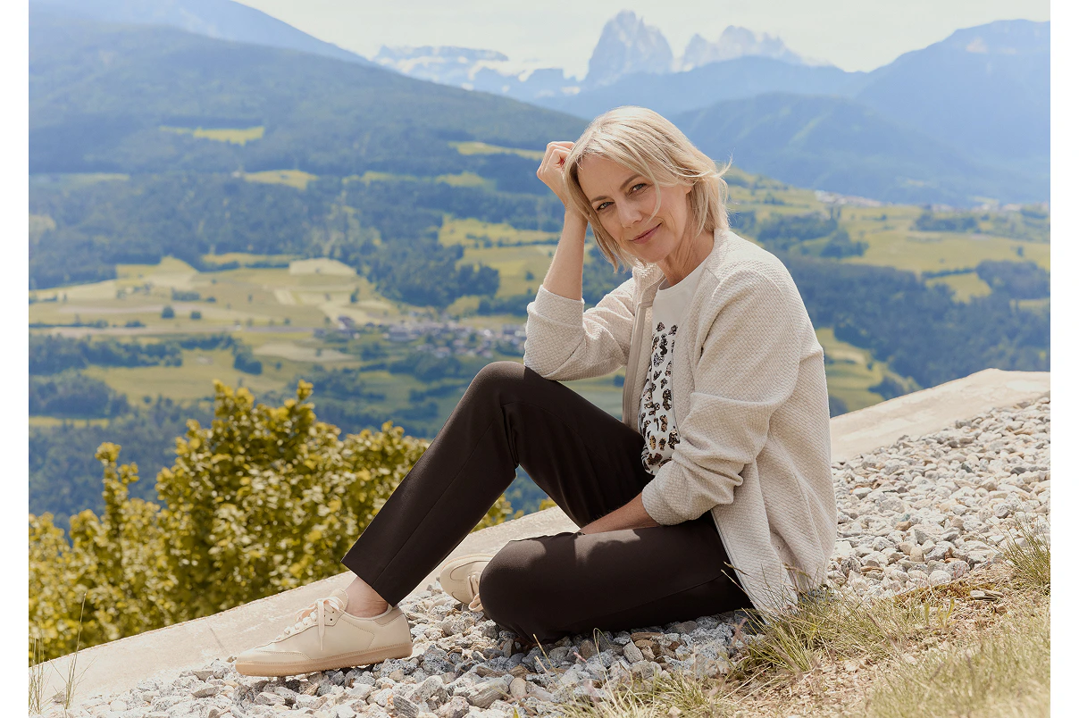 Een vrouw die zit, gekleed in een bruine broek, een shirt met patroon en een beige sweaterjasje. 