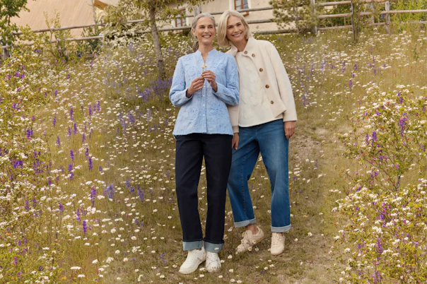 Zwei Frauen stehend in blauer Jeans, blauer Bluse und beiger Jacke. 