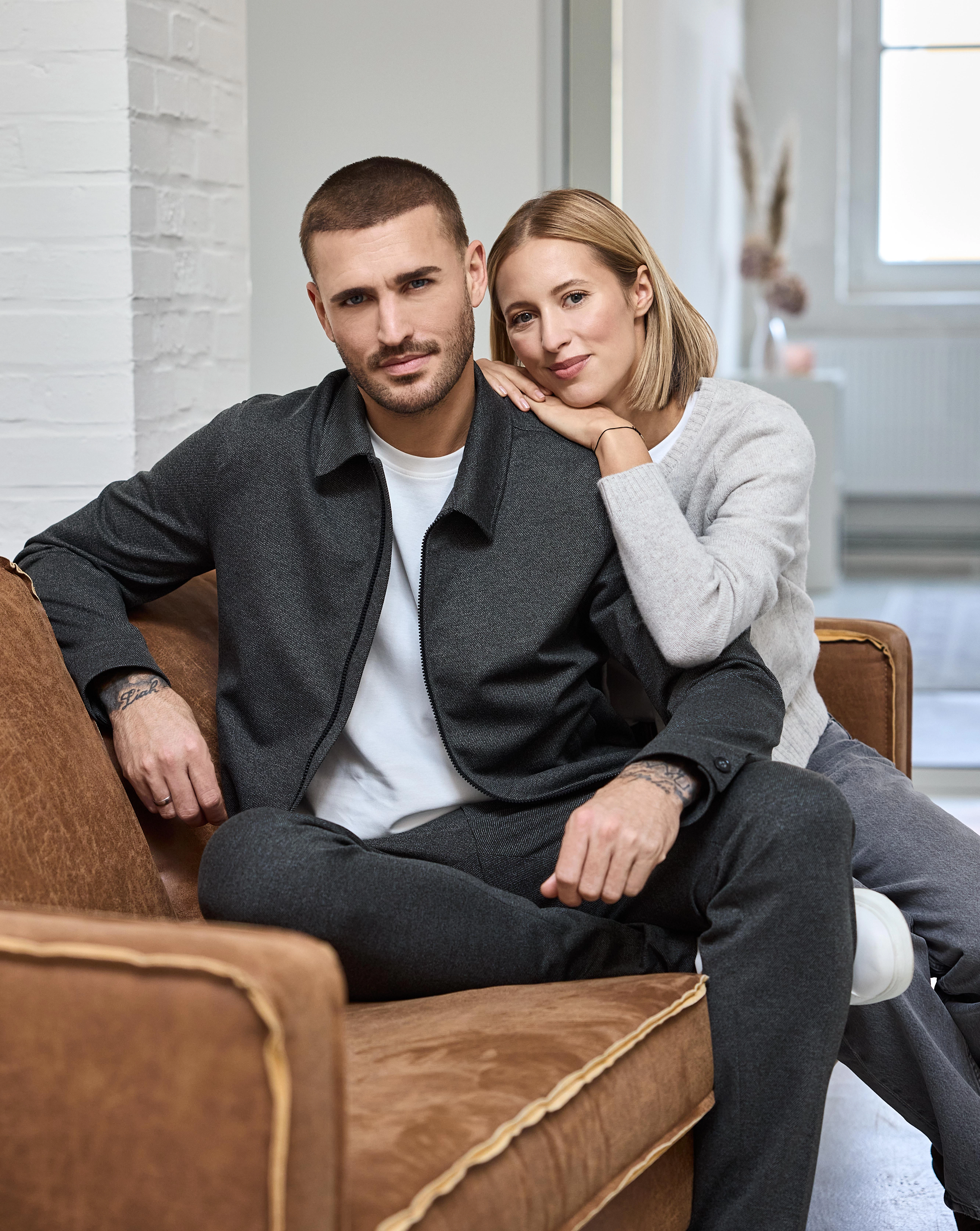 Ein Model trägt eine dunkelgraue Casual-Jacke über weißem T-Shirt, neben einer Frau im hellen Strickpullover auf einem Sofa.