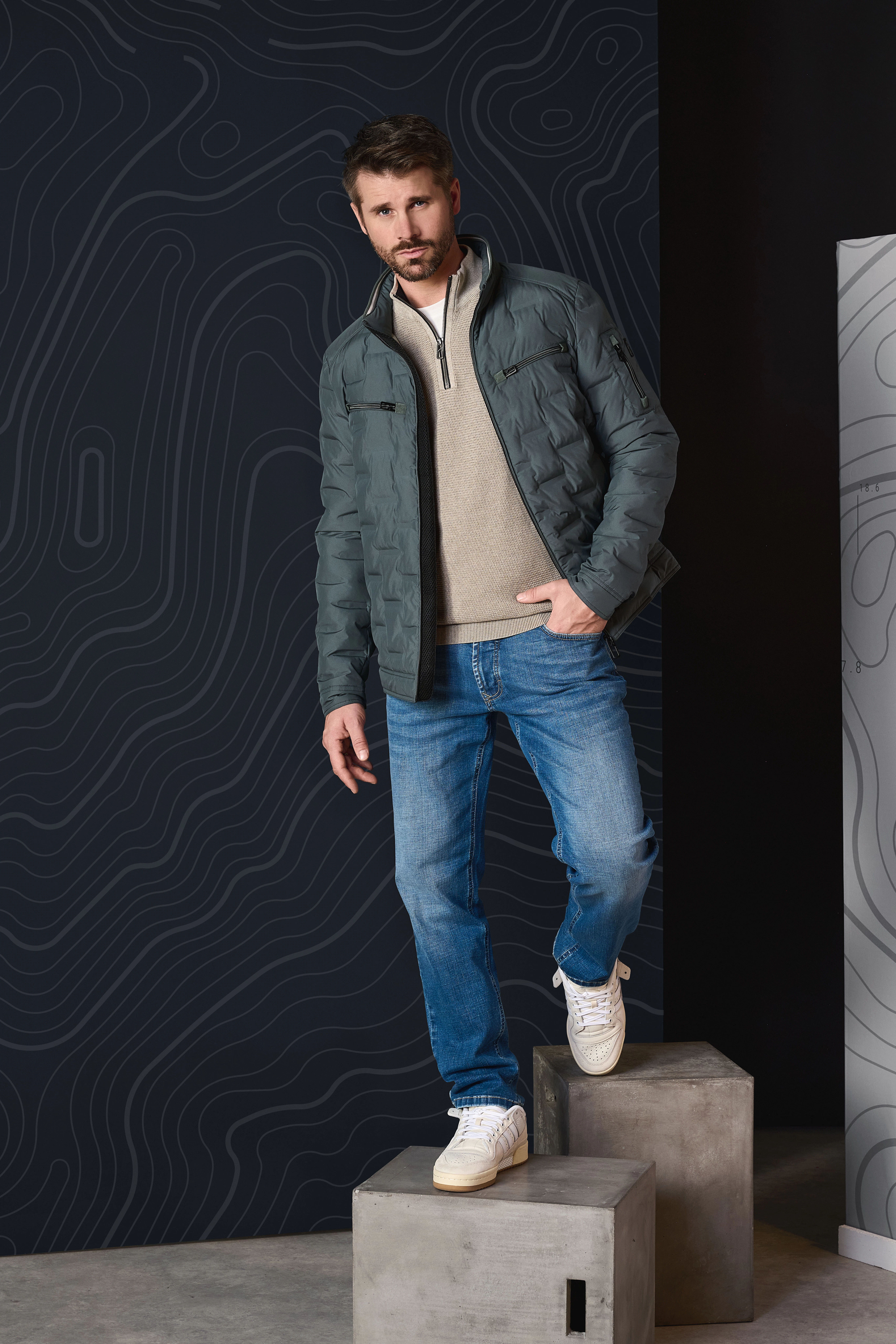 Thore Schölermann trägt eine dunkelgrüne Steppjacke über beigem Troyer, kombiniert mit blauer Jeans und weißen Sneakern.