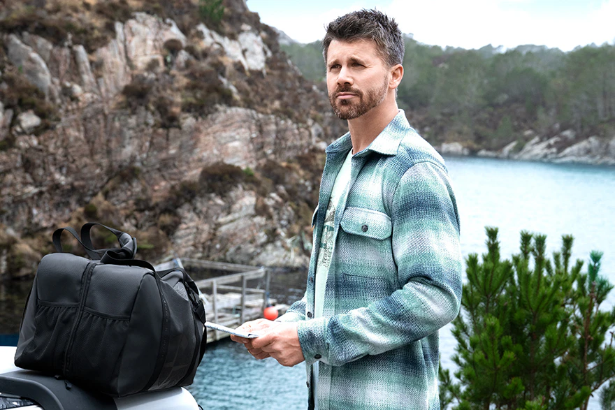 Thore Schölermann trägt ein grün-blau kariertes Overshirt über hellem T-Shirt vor einer felsigen Küstenlandschaft.