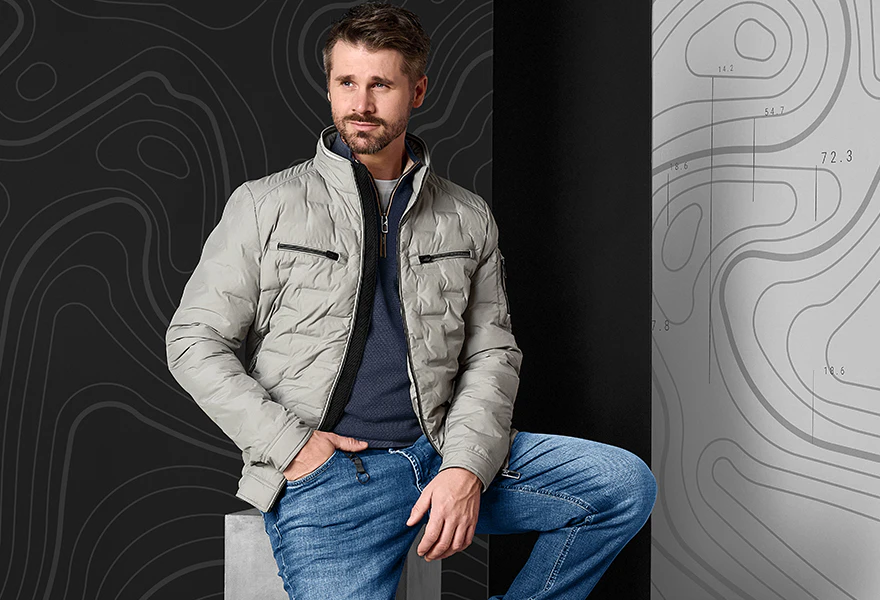 Thore trägt eine beige Herrenjacke mit Stehkragen, blauem Pullover und Jeans.
