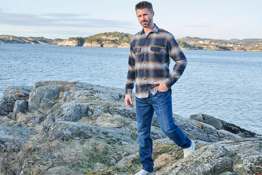 Thore Schölermann trägt ein blau-beige kariertes Overshirt mit blauer Jeans und weißen Sneakern an der Küste.