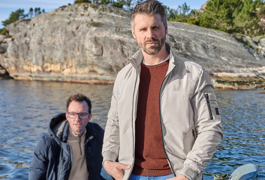 Thore Schölermann trägt eine beige Jacke über rostbraunem Pullover und blauer Jeans, begleitet von einem Mann im Boot.