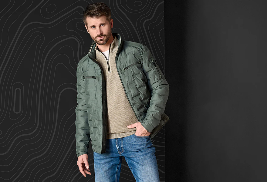 Thore trägt eine dunkelgrüne Herrenjacke mit beigem Pullover und blauer Jeans.