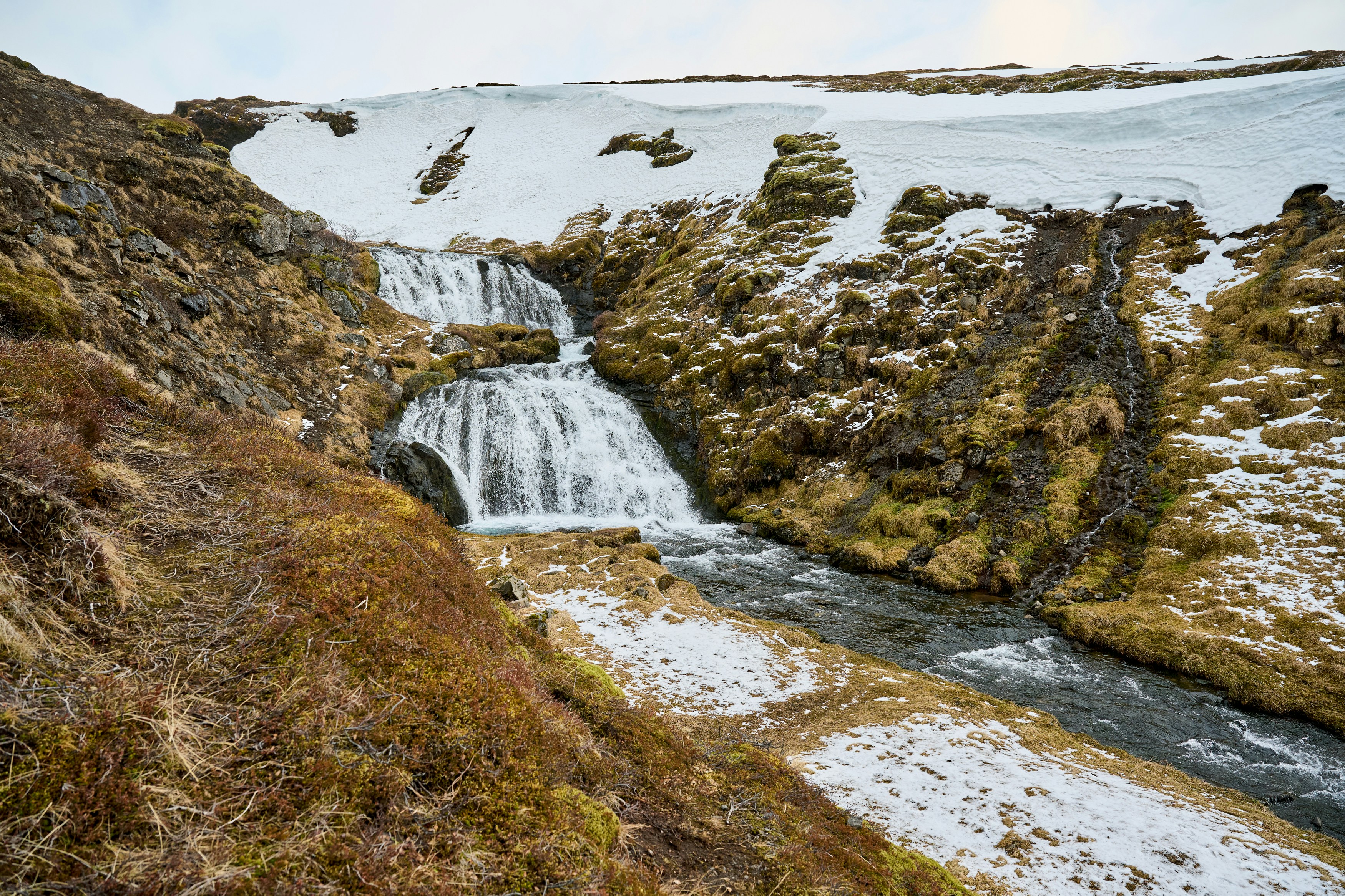 Kleiner Wasserfall mit Bach zwischen Schnee und moosigen Felsen