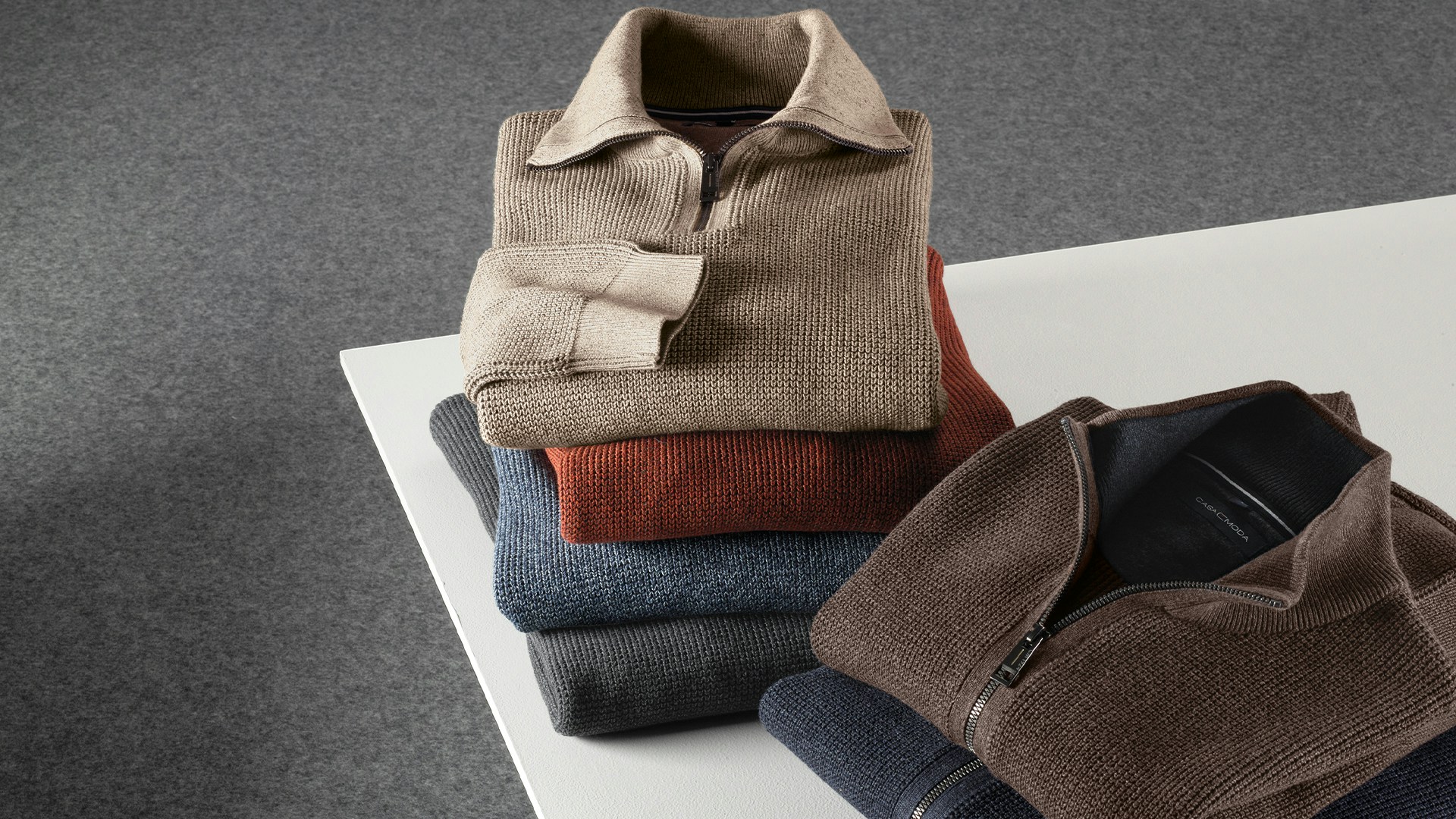 Fünf gefaltete Casamoda Troyer in Braun-, Blau- und Rosttönen auf hellem Tisch