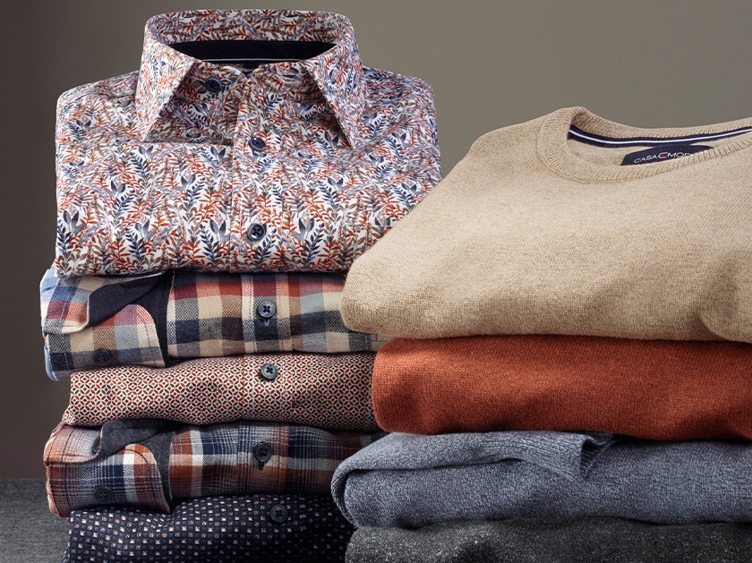Gefaltete Casamoda Hemden und Pullover in gedeckten Farben, gestapelt auf grauem Stoff