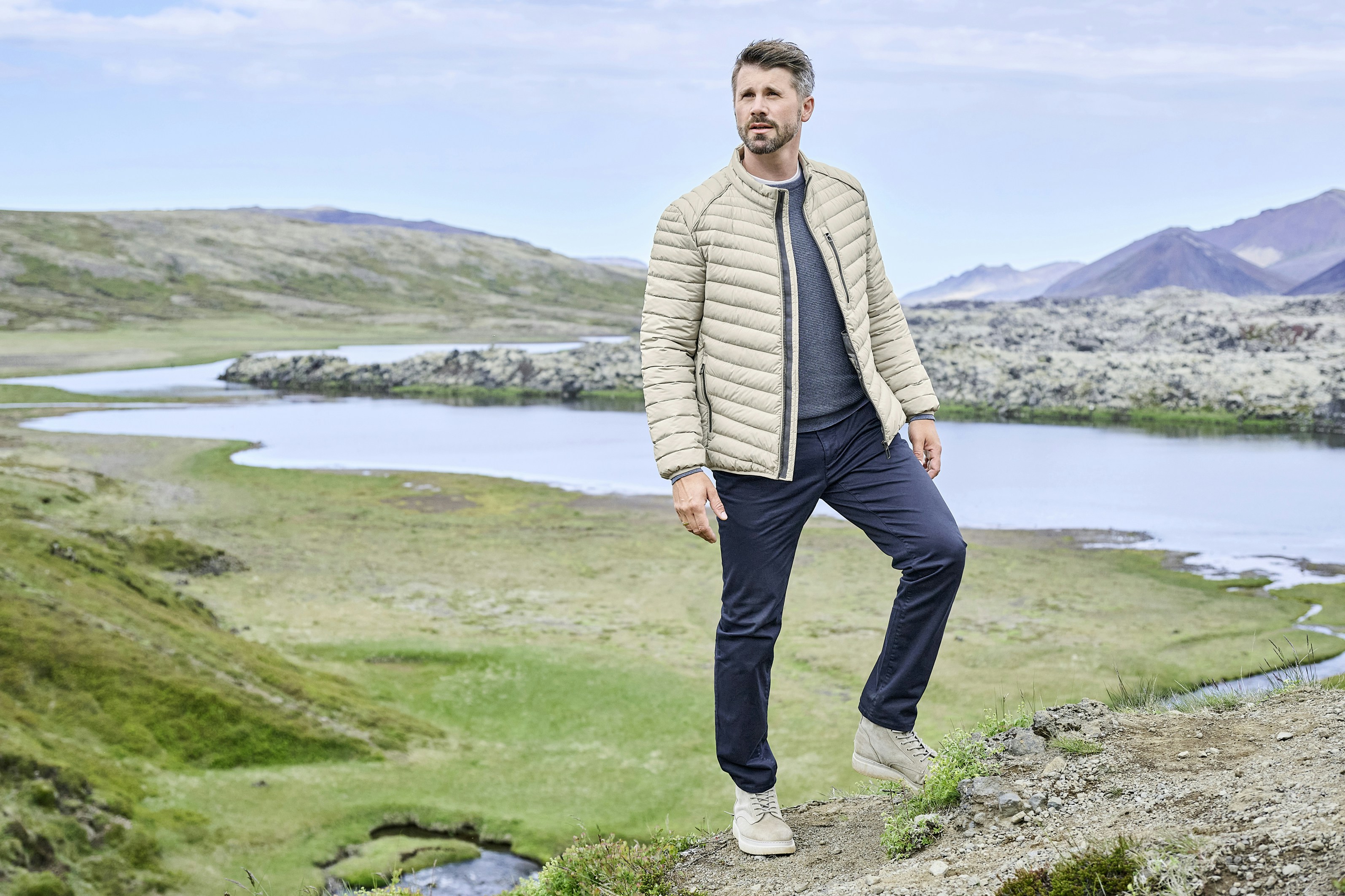 Thore Schölermann steht in hügeliger Landschaft mit See im Hintergrund und trägt eine beige gesteppte Jacke, dunkle Hose und helle Schuhe